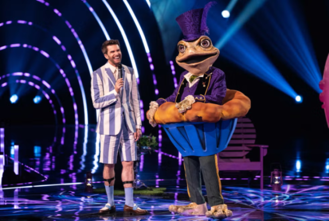 Comedian Jack Whitehall with a microphone next to a person in a frog costume, both on stage.