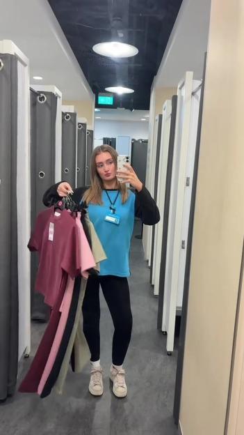 A woman holding several t-shirts on hangers in a changing room, taking a mirror selfie.