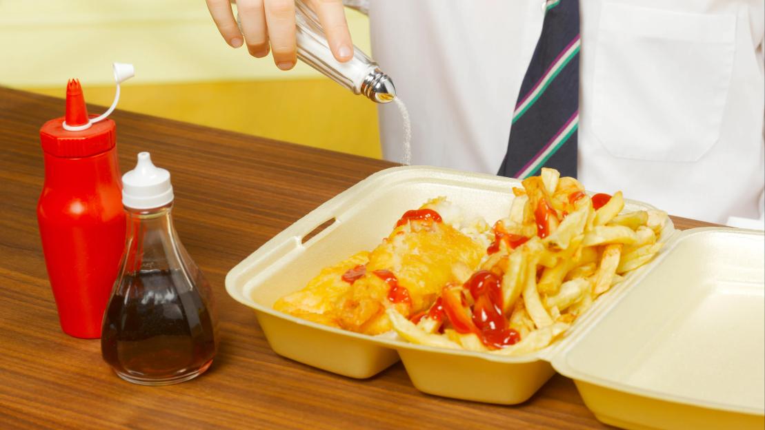 A boy pouring salt from a shaker onto fish and chips with ketchup in a polystyrene container.