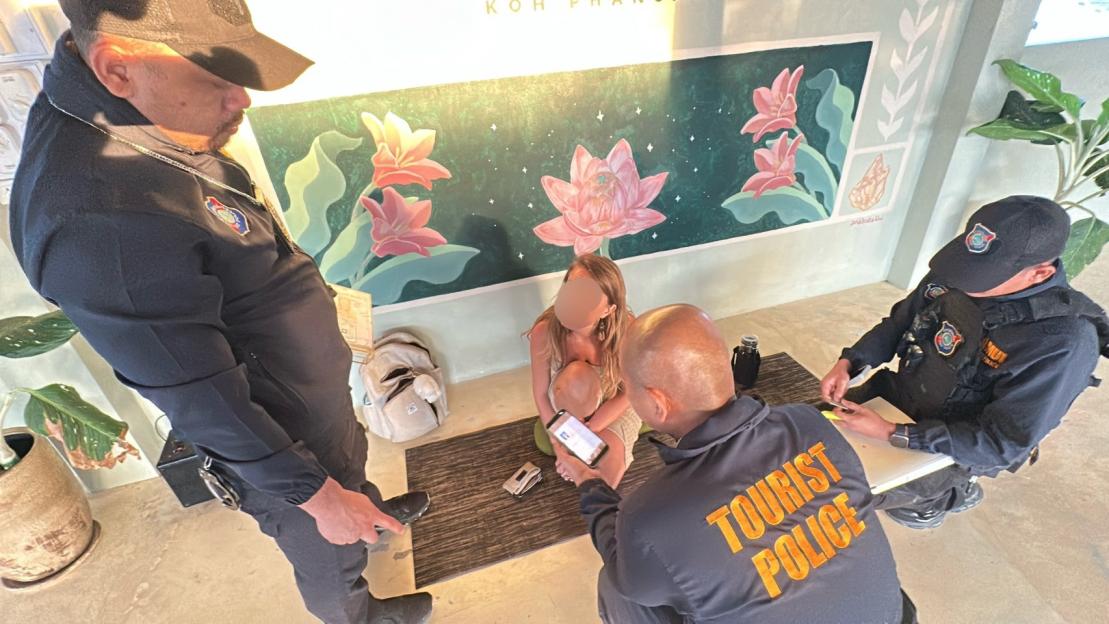 Two police officers questioning a woman, who is sitting on a mat, in front of a wall painting of flowers.