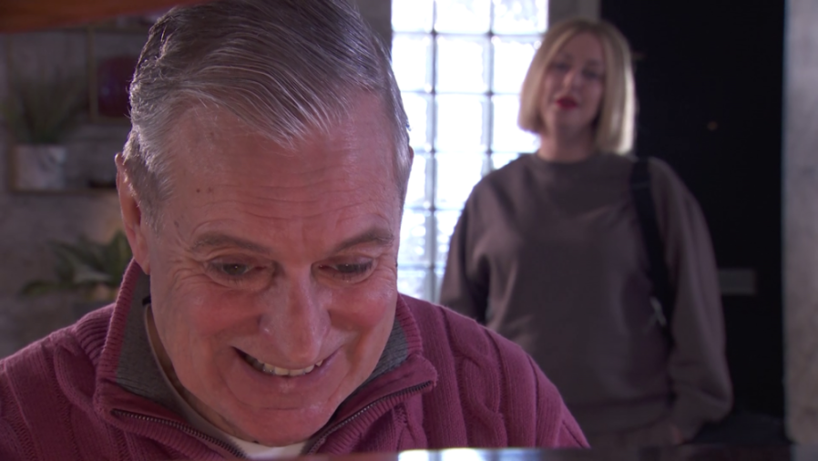 An elderly man in a pink sweater smiles as a blonde woman with red lipstick stands behind him.