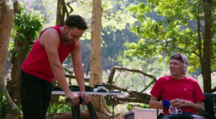A man in a red tank top stands, looking down at a table, while an older man in a red t-shirt and white bandana sits and looks towards the right.