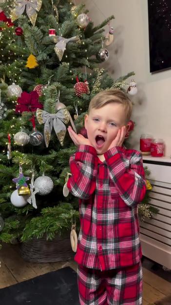 A child in red plaid pajamas standing in front of a decorated Christmas tree with hands cupped around his face, expressing excitement.