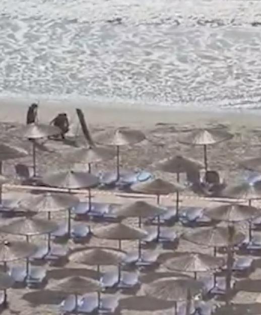 Bomb squad personnel pull an explosive-laden drone out of the sea on a beach lined with umbrellas.