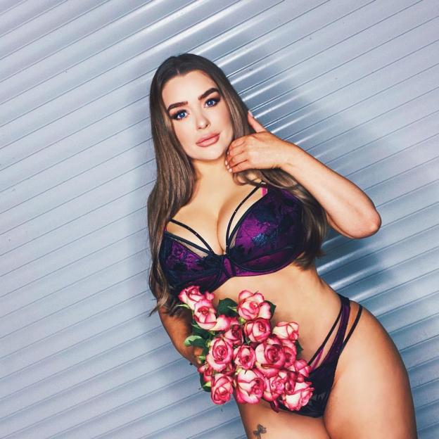 Amy Christophers posing in purple lingerie while holding a bouquet of roses.