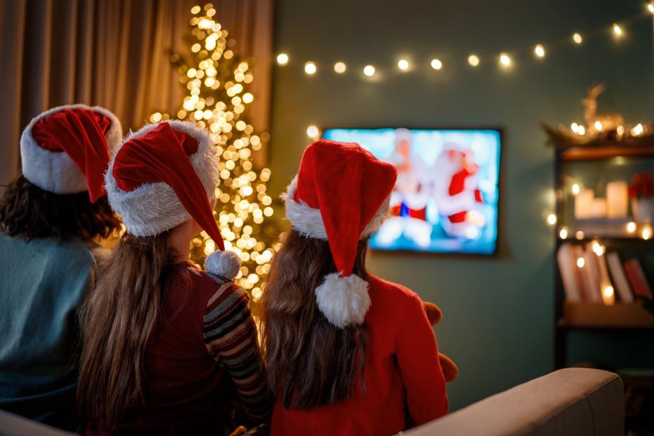 Family wearing Santa hats watching holiday movies on TV with a Christmas tree in the background.