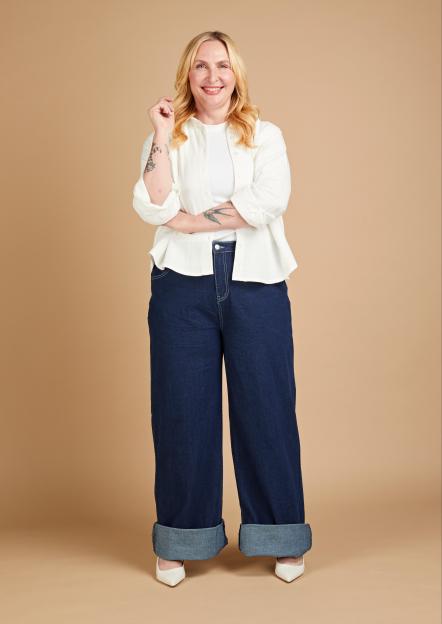 Annikki Sommerville wearing wide-leg, dark blue jeans with large cuffs, a white ribbed top, and an open white button-up shirt, posing against a light brown background.