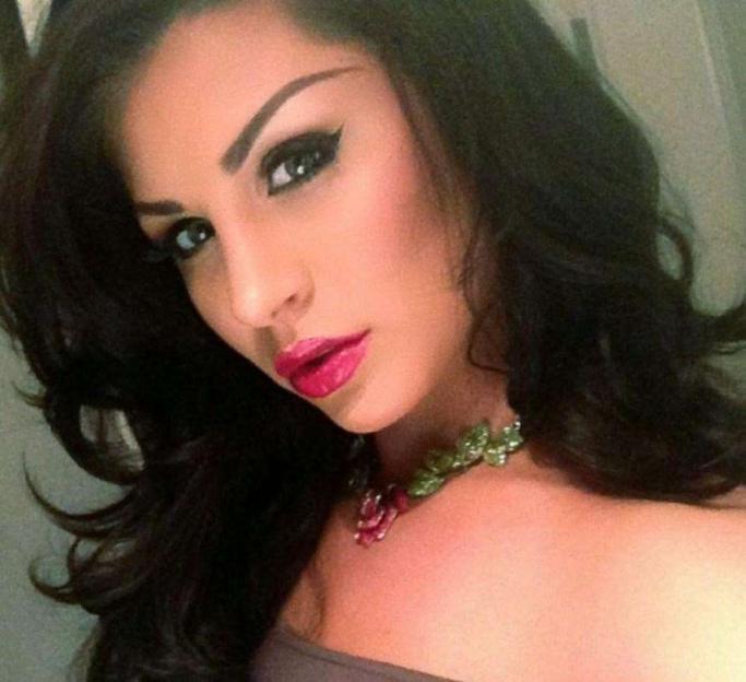 A woman with dark hair, dark eyeliner, and bright pink lipstick looks at the camera, wearing a necklace with green leaves and red flowers.