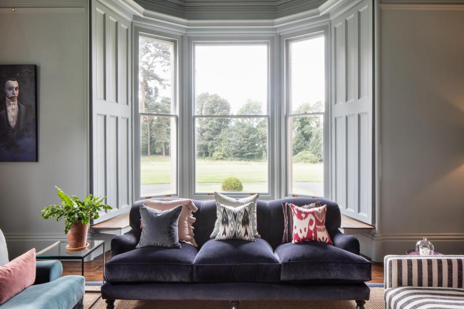 A navy blue velvet sofa with throw pillows in front of a bay window.
