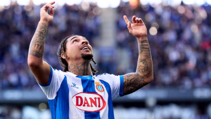 A soccer player with arm and neck tattoos looks up with arms raised.