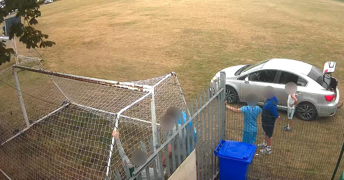 CCTV footage shows blurred figures of people and a car with its trunk open next to a soccer goal and a fence on a dry grassy field.