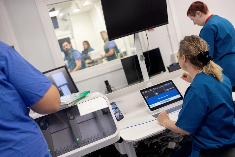 Surgeons in scrubs remotely operating robotic equipment with a tablet and screens, as other surgeons watch from behind a glass barrier.