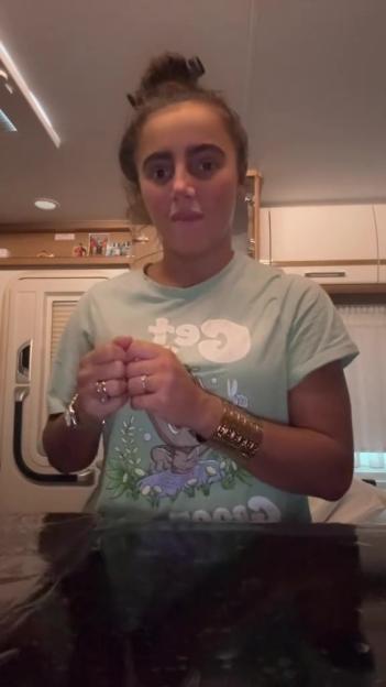 A woman wearing a light green t-shirt with a cartoon graphic on it, and several rings and bracelets, speaks to the camera indoors.