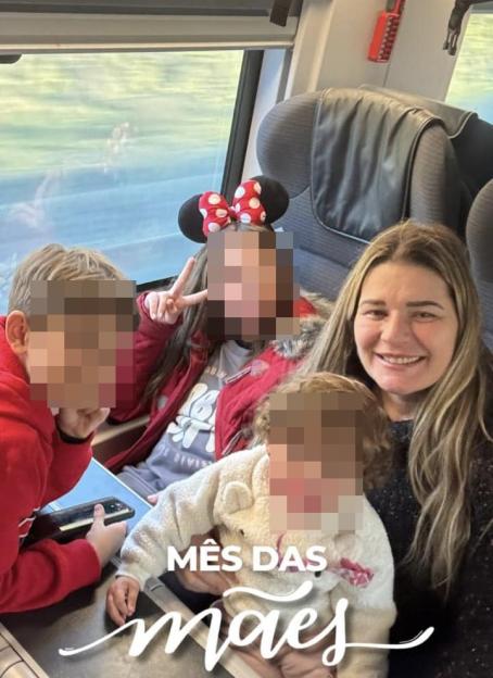 A smiling woman with long blonde hair sits with three children, one infant, one young girl making a peace sign, and one boy holding a phone.