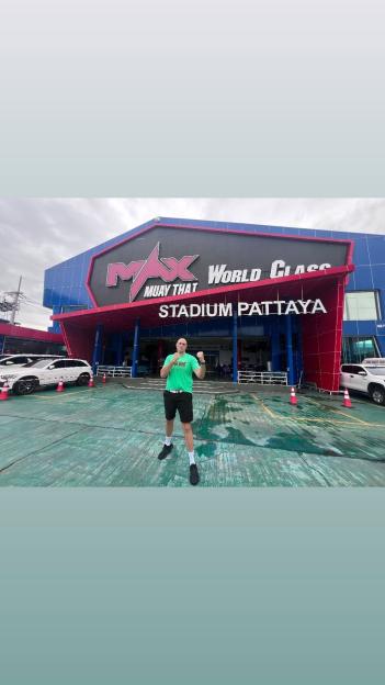 A man in a green t-shirt and shorts poses with fists up in front of the Max Muay Thai World Class Stadium Pattaya.