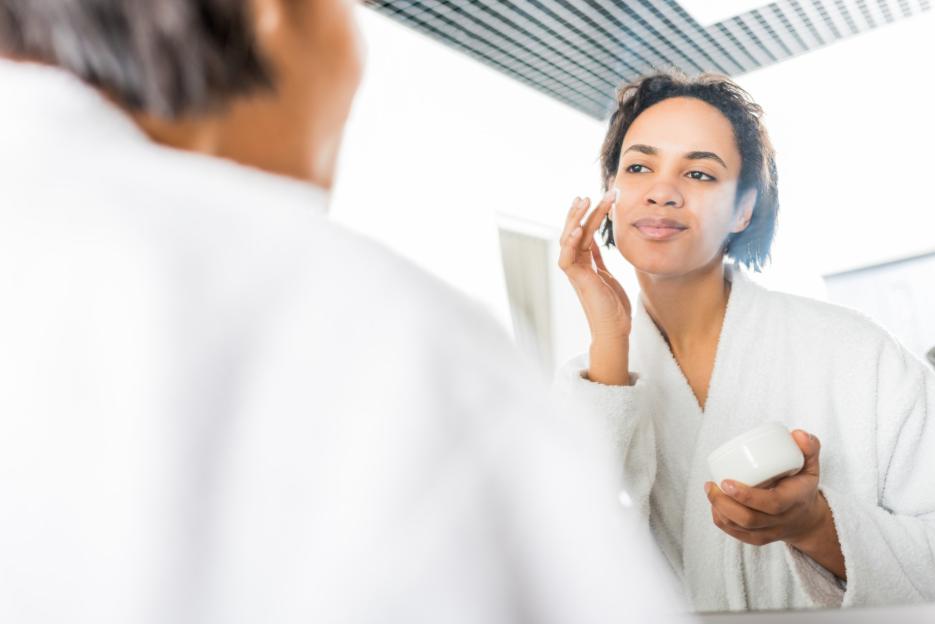 Woman in white robe applying moisturizer to her face while looking in a mirror.