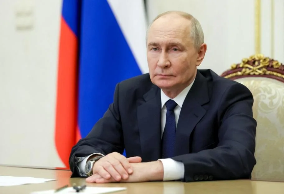 Russian President Vladimir Putin sits at a desk during a meeting in Moscow, with the Russian flag in the background.
