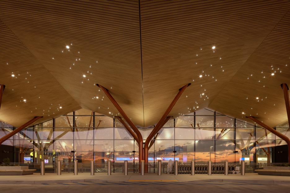 The exterior of an airport terminal with a wooden ceiling and orange supports, reflecting a sunset sky.