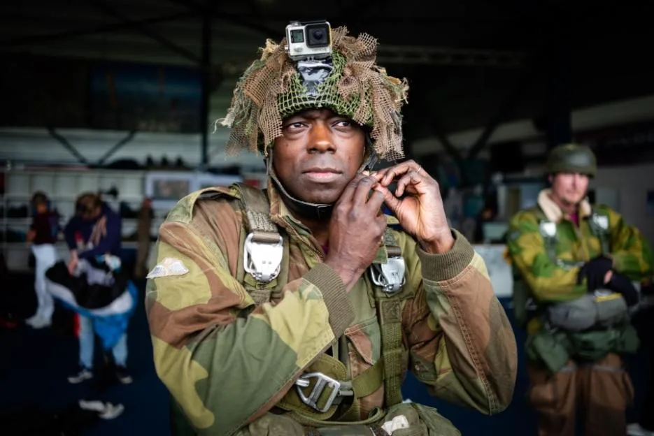 MP Bayo Alaba wearing a helmet with a GoPro camera and camouflage parachute gear, preparing to board a Dakota C-47.
