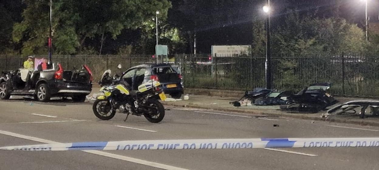 Police tape blocks off a road where a car has been heavily damaged and a police motorbike and another car are also visible.