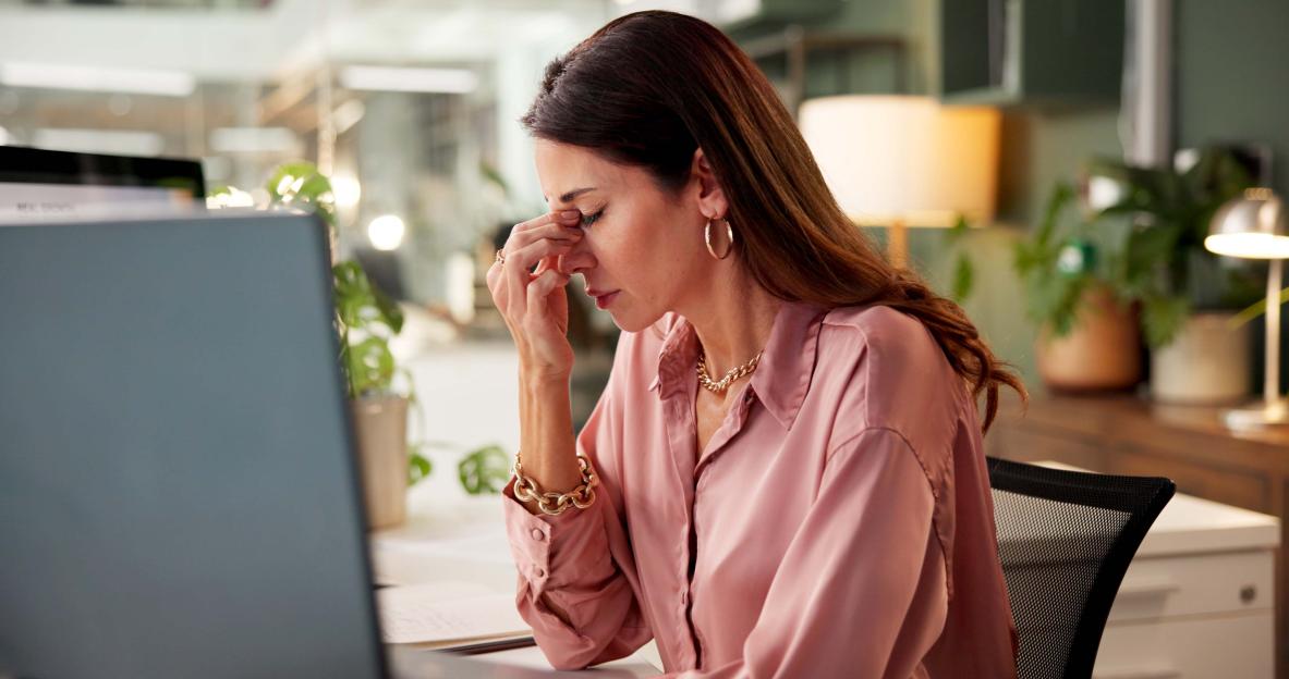 A businesswoman with a headache rubs her temples while sitting at a desk in an office at night.