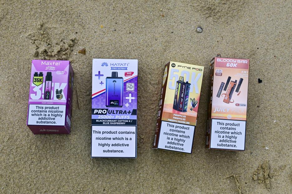 Four vape product boxes with nicotine warnings sit on sand.