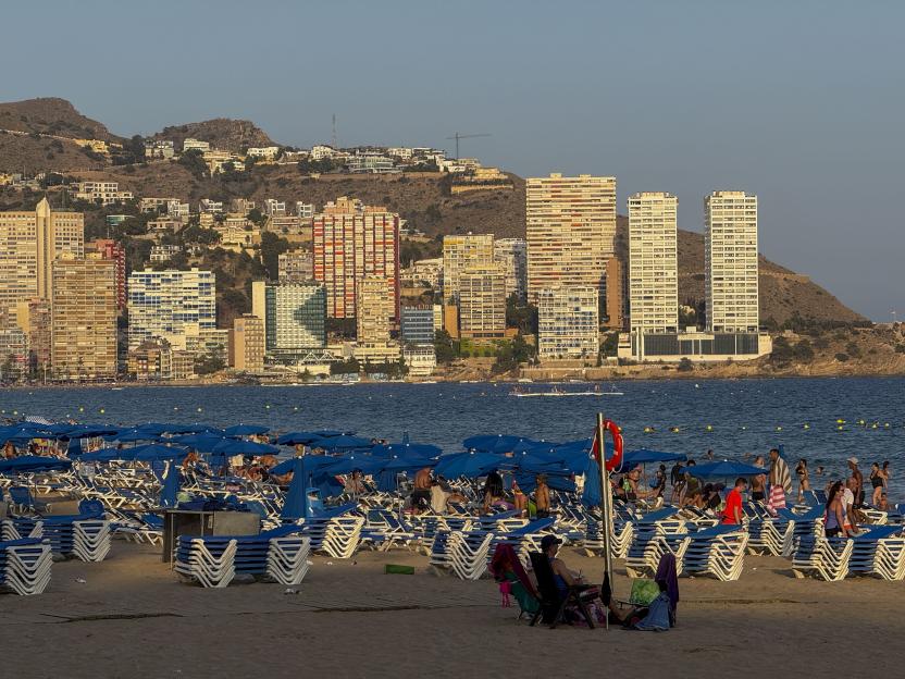 A crowded Benidorm beach with blue umbrellas and sun loungers, with high-rise buildings and mountains in the background.