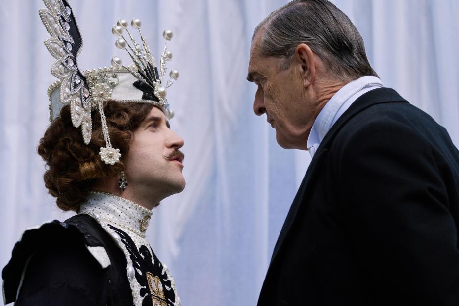 A person in an ornate, jeweled hat with a curled mustache faces a stern older man in a suit, both looking at each other intently.