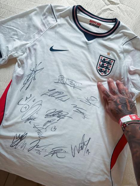 A white England football jersey signed by players, with a tattooed hand pointing at the signatures.