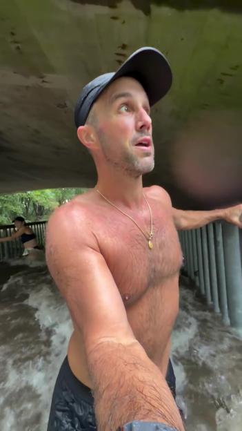 A man in a baseball cap stands shirtless in rapidly moving water under a bridge, looking up.