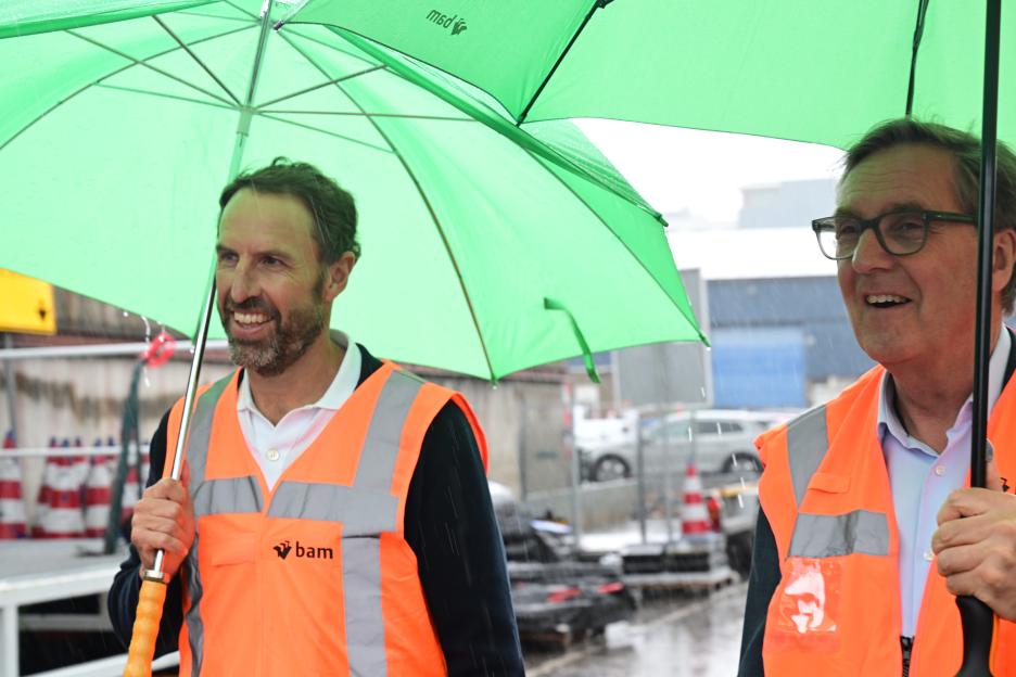 Gareth Southgate and Alan Milburn in orange safety vests holding umbrellas in the rain.