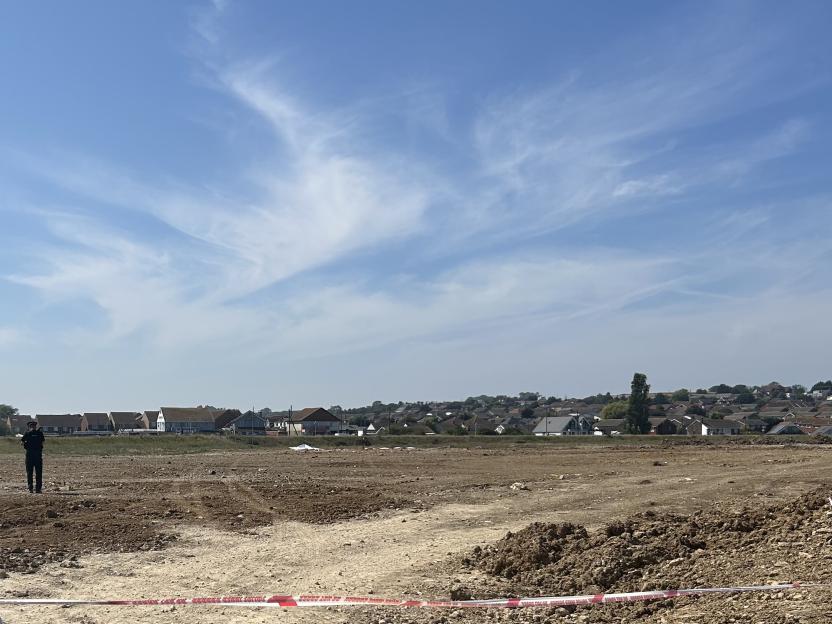 Police officer on a dirt field with a crime scene cordon, with houses and blue sky in the background.