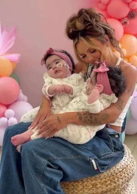 Jesy Nelson holds her twin daughters, Ocean and Story, on their first birthday in front of a pink balloon display.