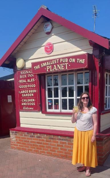 A woman standing in front of the Signal Box Inn, described as "THE SMALLEST PUB ON THE PLANET".
