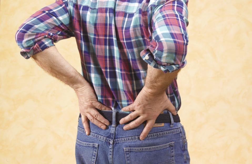 A senior man in a plaid shirt and jeans holds his lower back in pain.