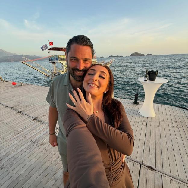 A smiling couple on a dock at sunset, the woman showing off an engagement ring.