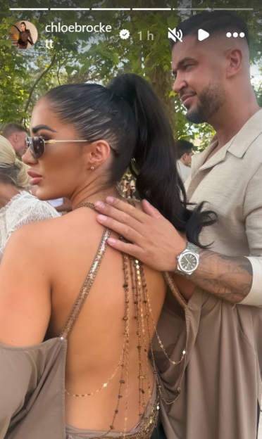 A man with a watch on his wrist places his hand on the bare back of a woman wearing a backless dress and sunglasses.