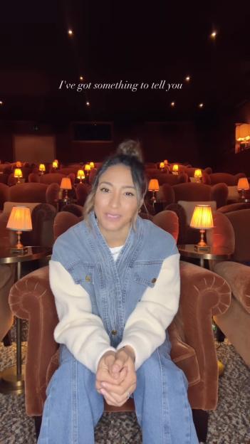 Karen Hauer, wearing a denim jacket and jeans, sits in a plush chair within a dimly lit theater setting, with the text "I've got something to tell you" displayed above her.