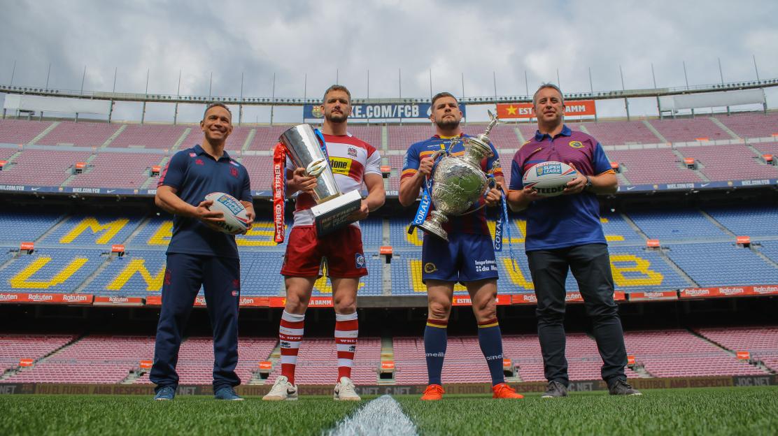 Two rugby players holding trophies stand between two men holding rugby balls on a football pitch.