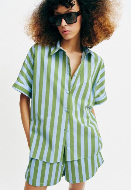 A person wearing a striped co-ord set featuring a short-sleeved button-up shirt and shorts, in alternating vertical stripes of light blue and olive green.