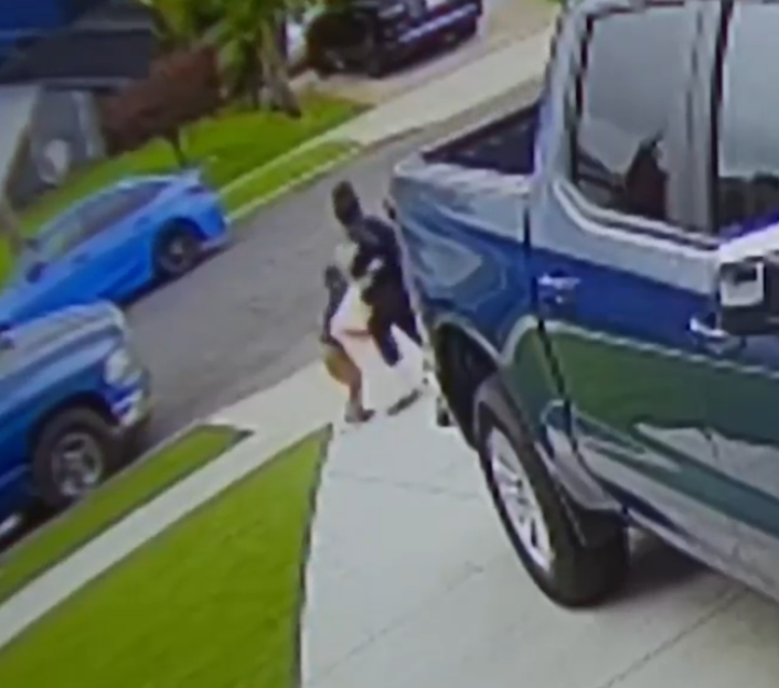 A person in dark clothing grappling with another person on a sidewalk next to a blue pickup truck.