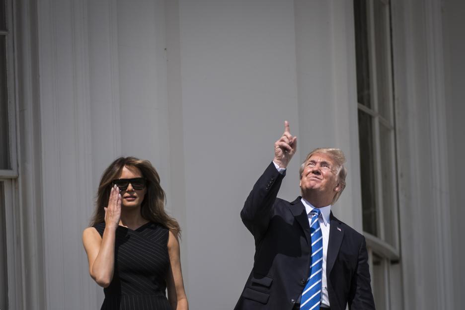 Donald Trump pointing at the solar eclipse without protective glasses, with Melania Trump shading her eyes beside him.