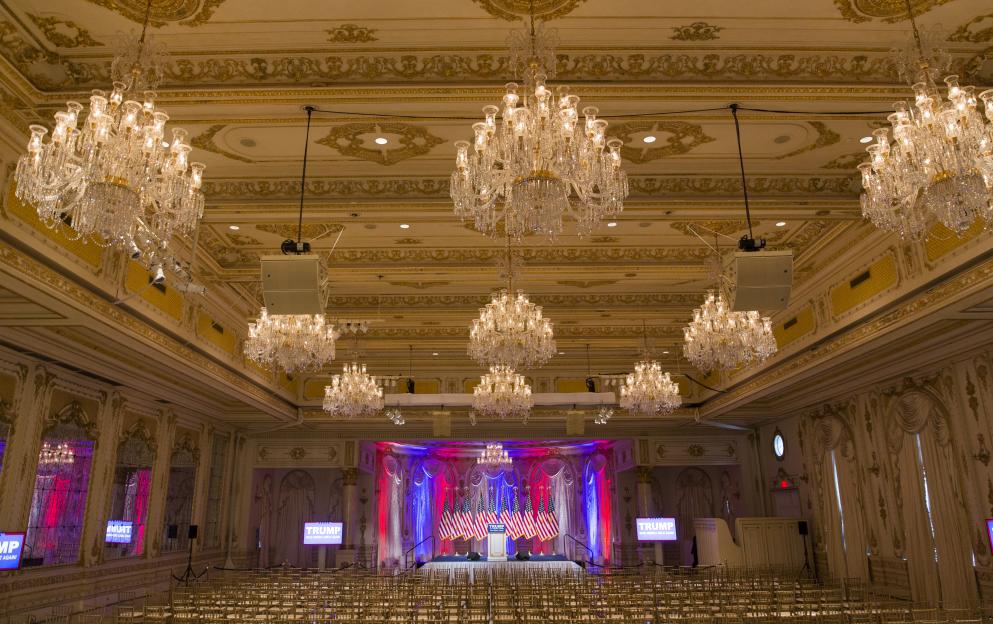 The Donald J. Trump Ballroom at the Mar-A-Lago Club, set up for a rally with a podium, American flags, and a "TRUMP MAKE AMERICA GREAT AGAIN" sign.