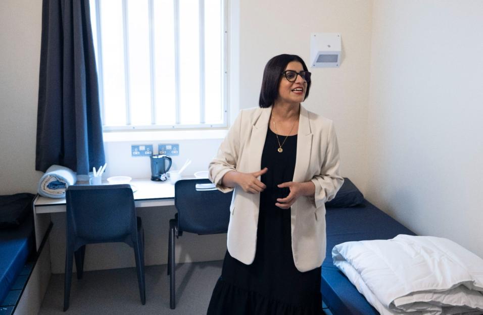 A woman in a black dress and cream blazer stands in a small room with two beds and a desk, similar to a prison cell.