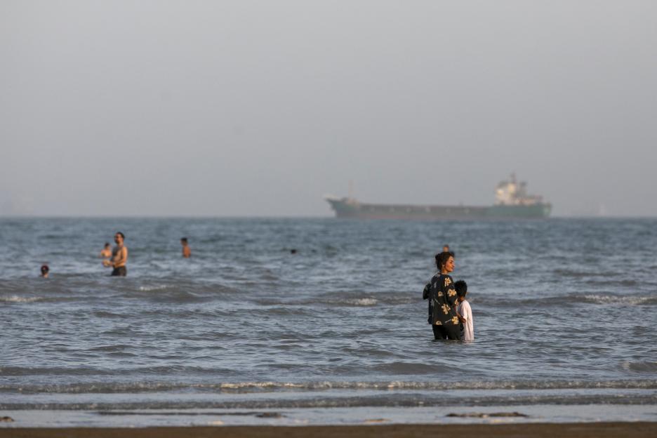 People swimming along the coast of the Persian Gulf in Bandar Abbas, Iran, with cargo vessels visible in the background.