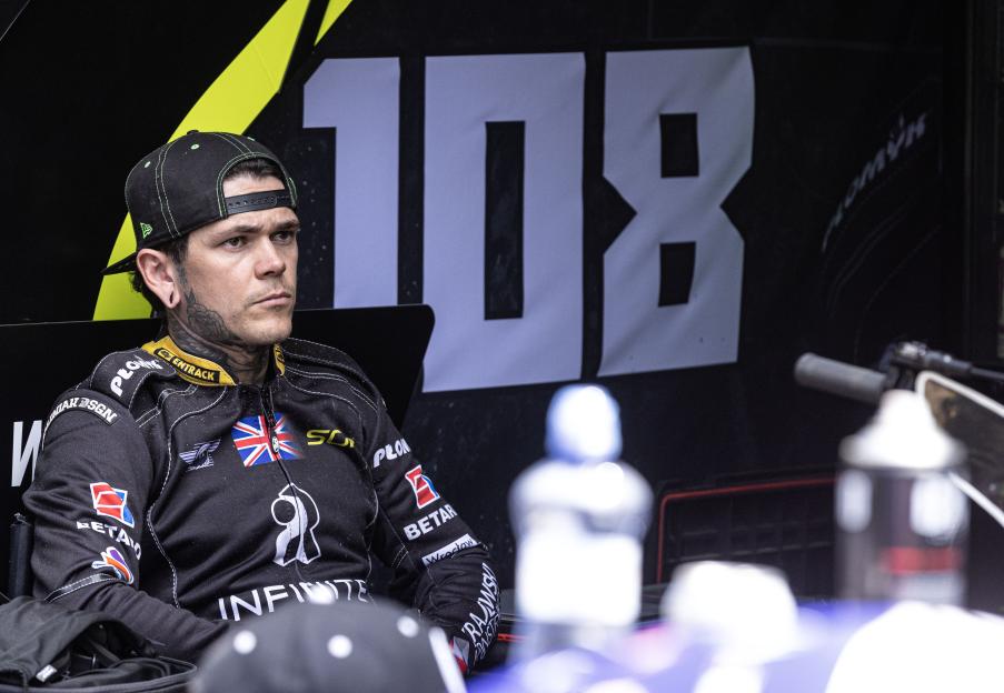 A man wearing a black cap and racing suit with a Union Jack flag on the shoulder, looking intently to the right, against a black background with a large white "108".