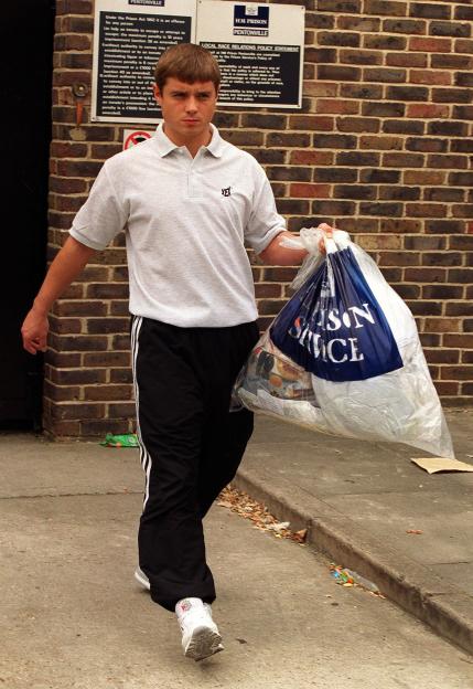 A man in a polo shirt and athletic pants walks away from a brick wall with prison notices, carrying a large plastic bag labeled "PENTONVILLE PRISON SERVICE".