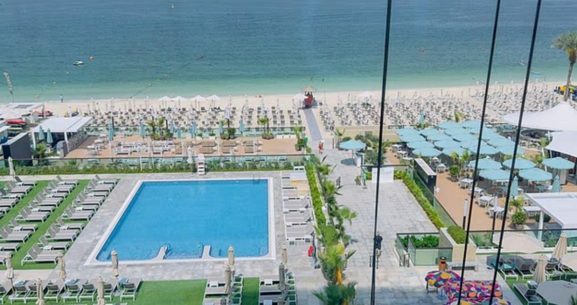 An outdoor pool next to a sandy beach with many lounge chairs and blue umbrellas.