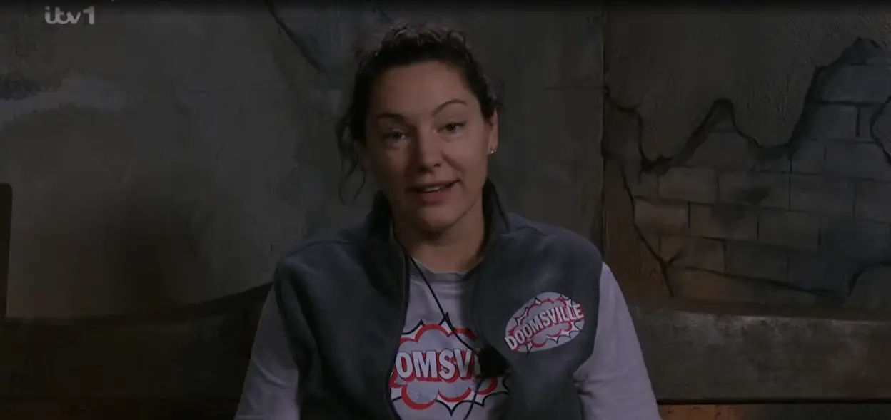 A woman speaking, dressed in a grey vest over a white t-shirt that says "DOOMSVILLE."