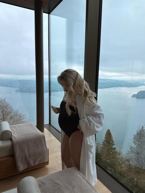 Molly-Mae Hague in a black swimsuit and white robe with a baby bump, standing by a window overlooking a lake and mountains.